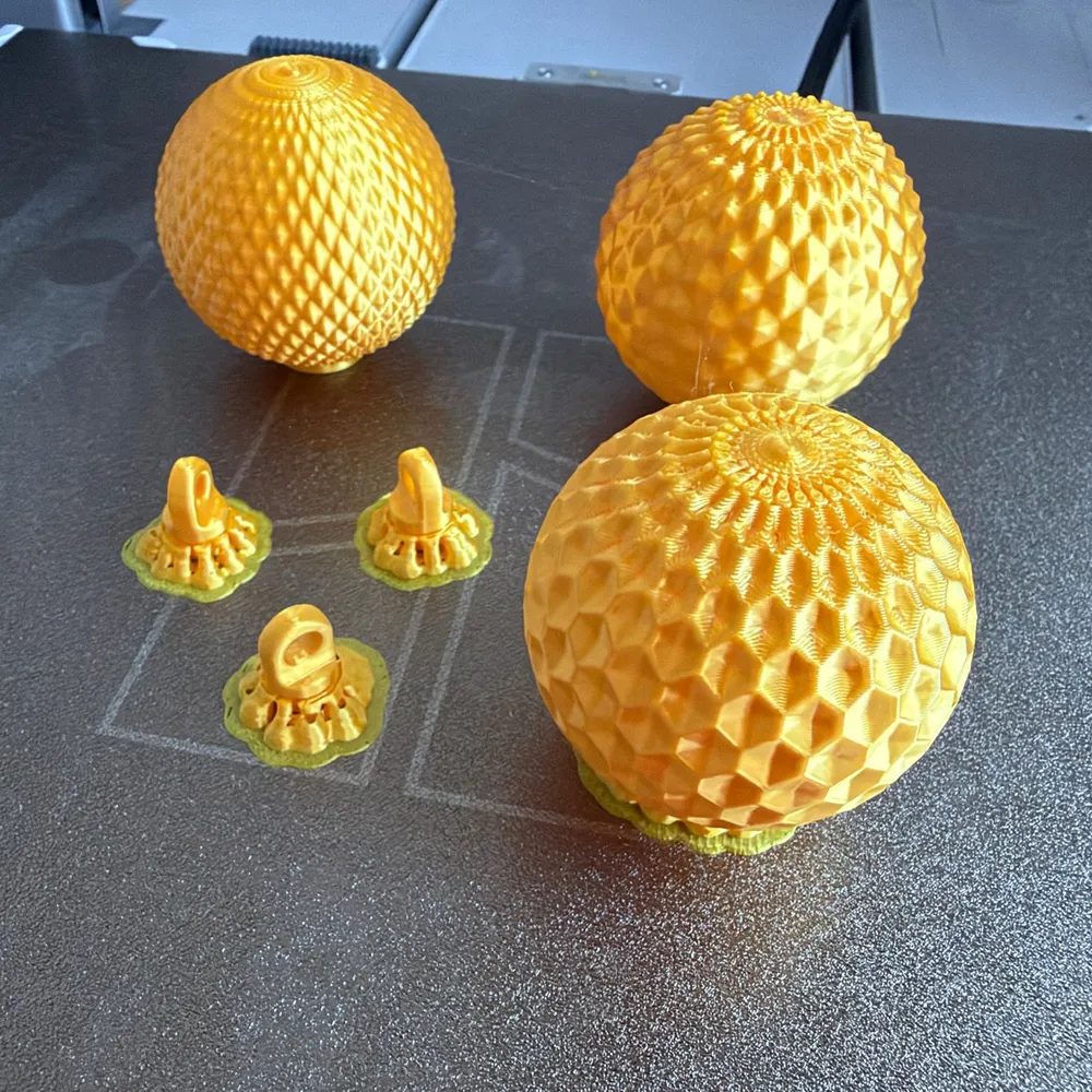 Tres boles de Nadal impreses en 3D en filament daurat, amb patrons calats diferents, i tres penjadors sobre els suports d'impressió